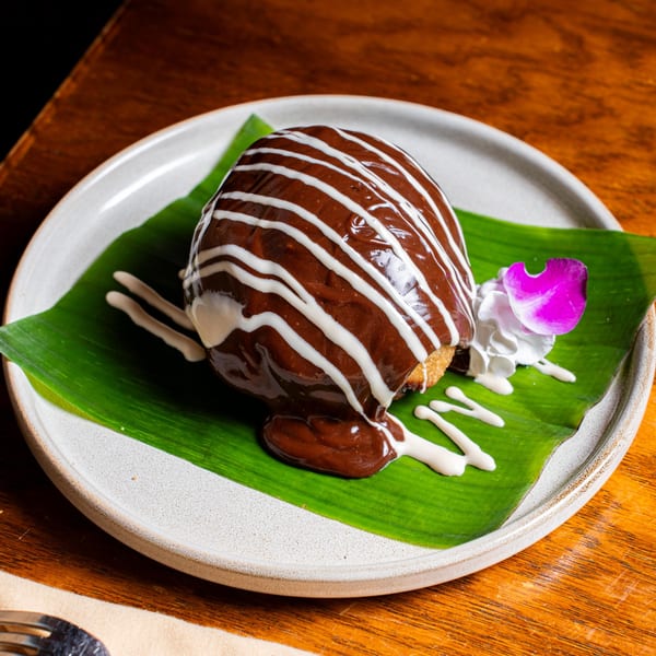 Nur Thai dessert: a chocolate-covered pastry with white drizzle, whipped cream, and a purple orchid on a banana leaf.