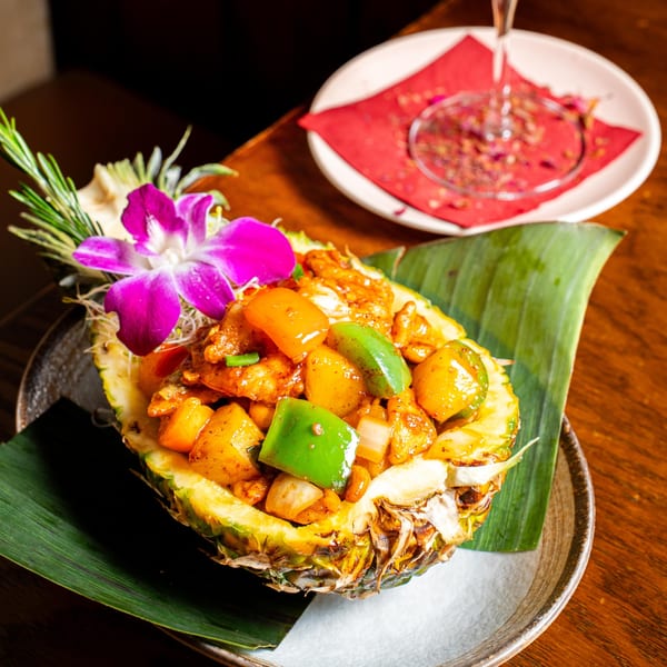 Sweet and sour chicken with pineapple and bell peppers served in a pineapple half, garnished with an orchid.