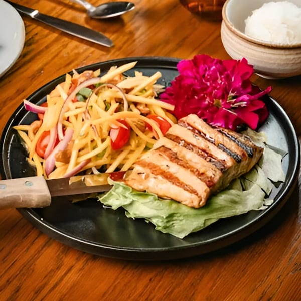 Grilled fish with mango salad and a pink flower garnish on a black plate at Nur Thai.