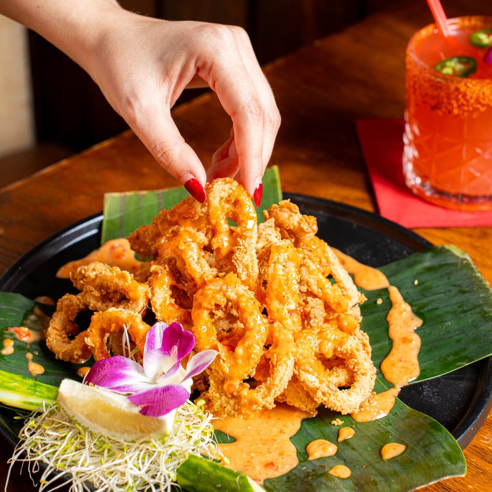 A hand picks up a piece of crispy calamari from a plate with sauce, lime, sprouts, and a drink.